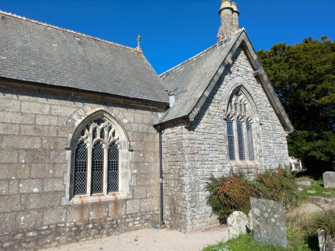 Side view of a historic stone church with Gothic-style windows and a slate roof, surrounded by greenery.