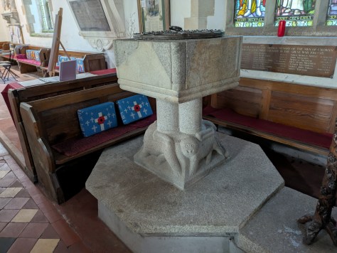 A stone baptismal font with a decorated base featuring animal carvings, set in a church interior with wooden pews and stained glass windows in the background.